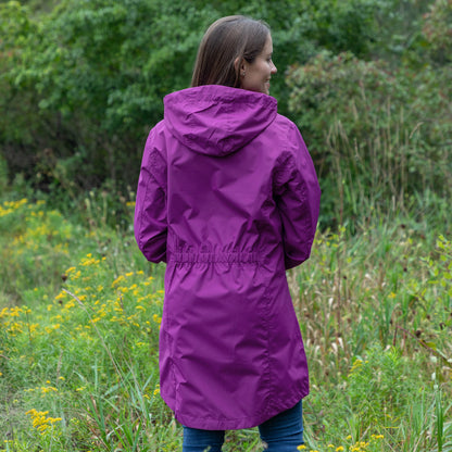 A woman wearing a purple hooded raincoat is photographed from behind in an outdoor grassy field with green foliage and yellow wildflowers in the background.