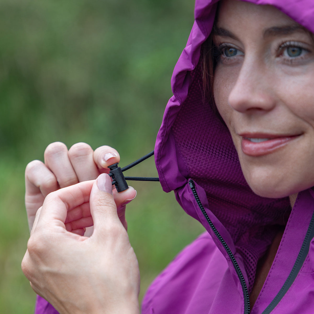 A close-up lifestyle shot of a woman wearing a purple hooded rain jacket, holding a small black plastic toggle with a black cord to demonstrate the hood's drawstring closure.
