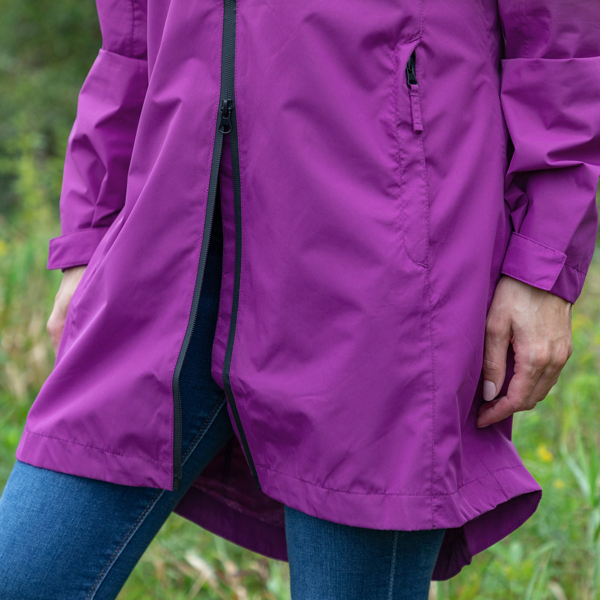 A person wearing a mid-length purple hooded jacket with a front zipper and zip pocket, paired with blue jeans, photographed outdoors against a blurred green grassy background.