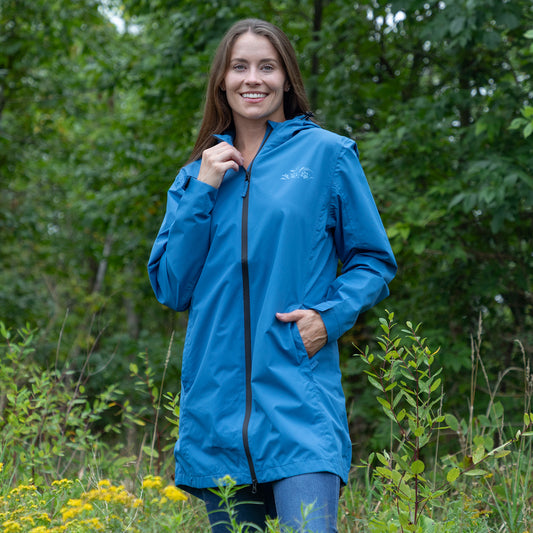 A woman wearing a bright blue hooded rain jacket with a black zipper, standing outdoors in a grassy area with green trees in the background.