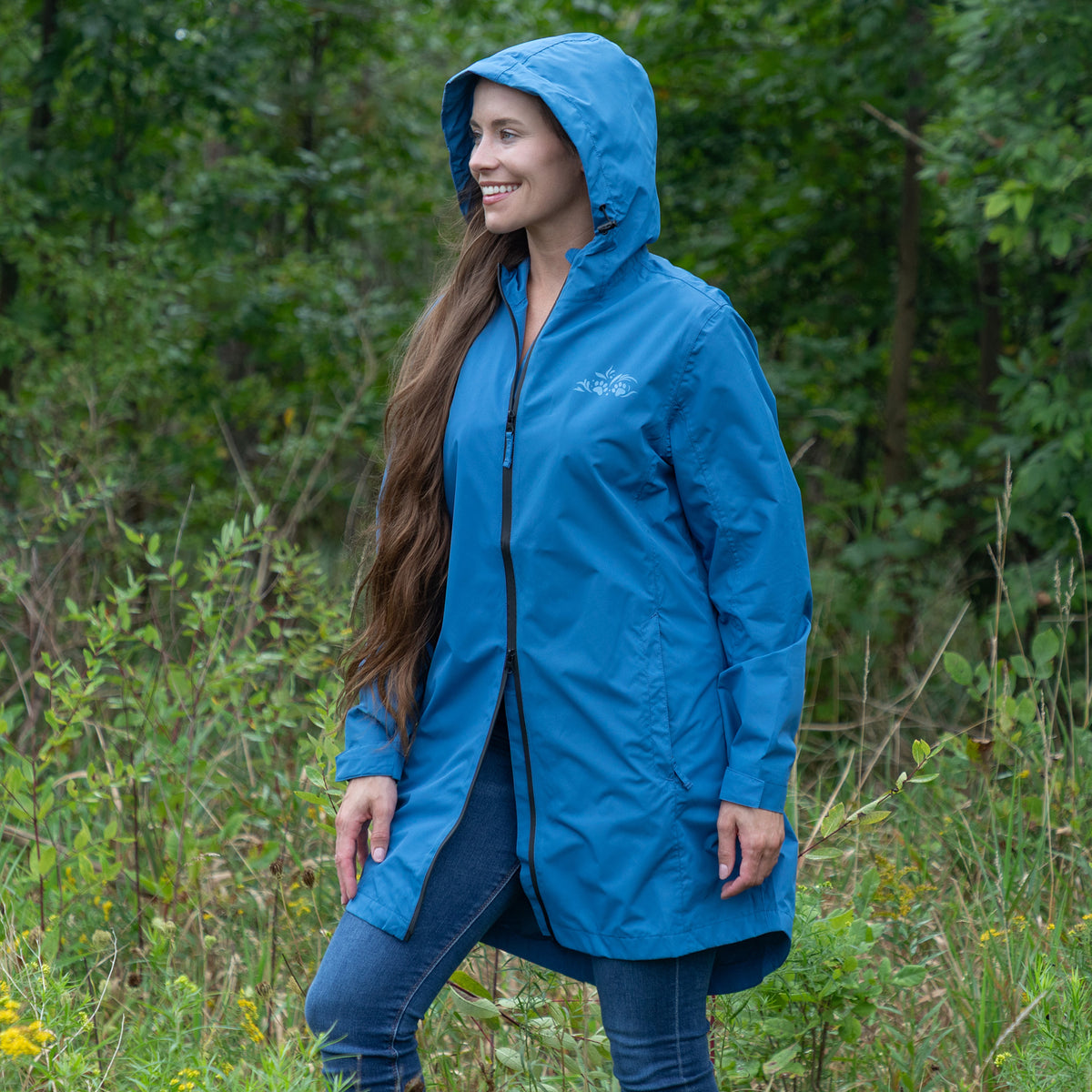 A woman wearing a bright blue hooded rain jacket stands outdoors in a grassy, wooded area. The jacket has a front zipper, long sleeves, and a small white logo on the left chest.