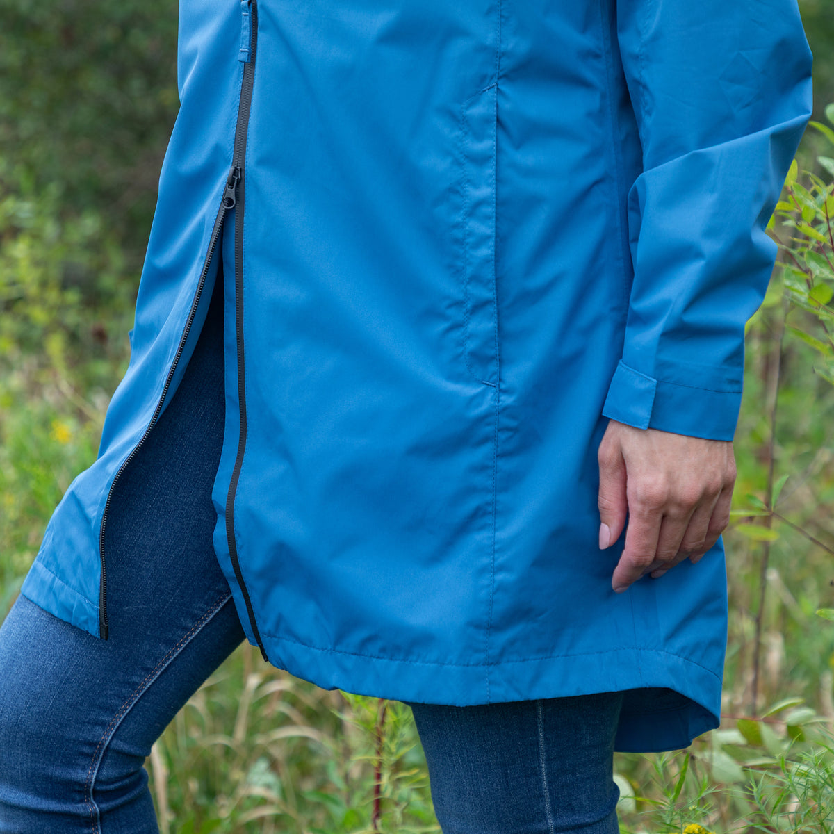 A person wearing a bright blue hooded jacket with a side zipper and blue jeans, standing outdoors in a grassy area.