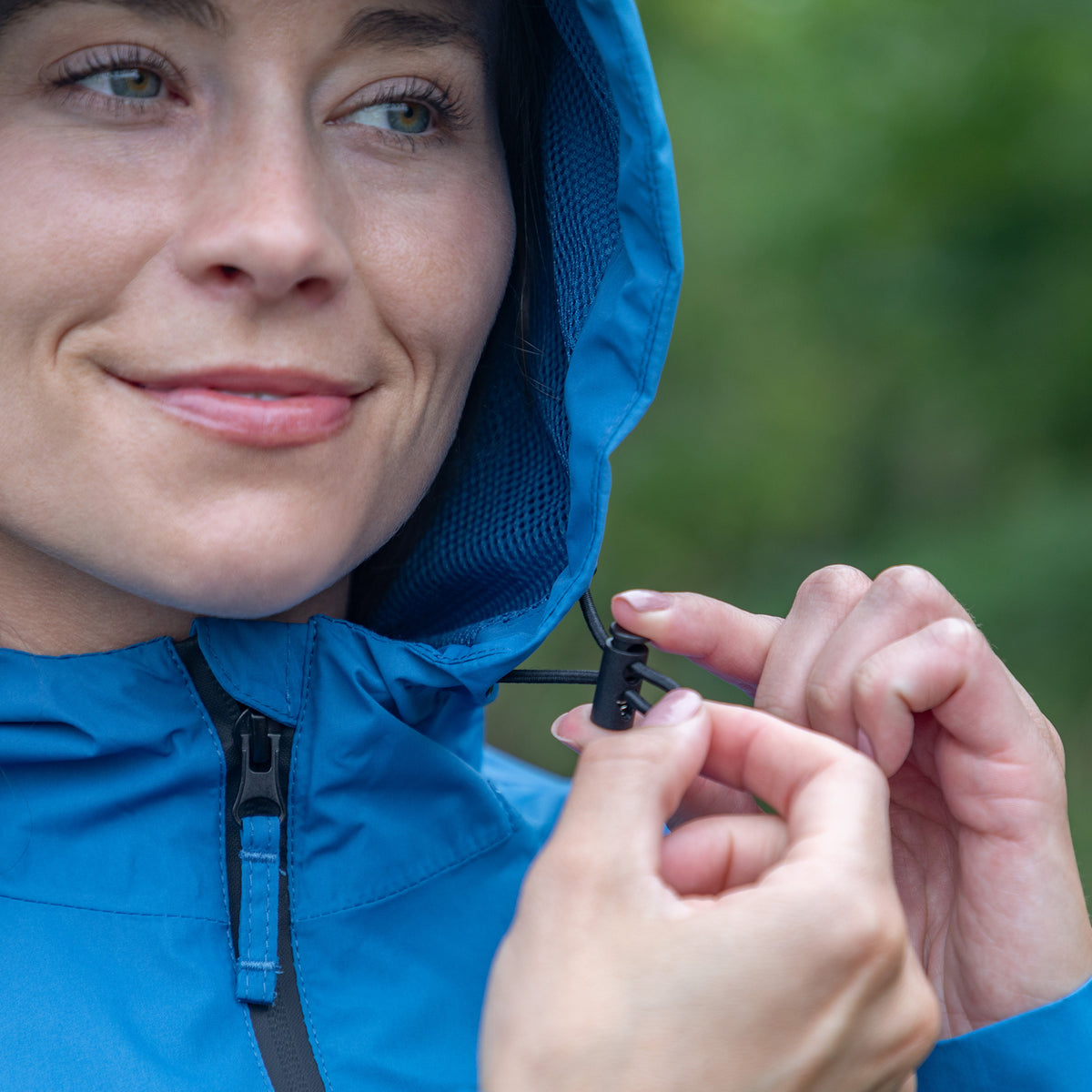 Close-up of a woman wearing a bright blue hooded jacket and adjusting the hood closure with her hands, set against a blurred green outdoor background.