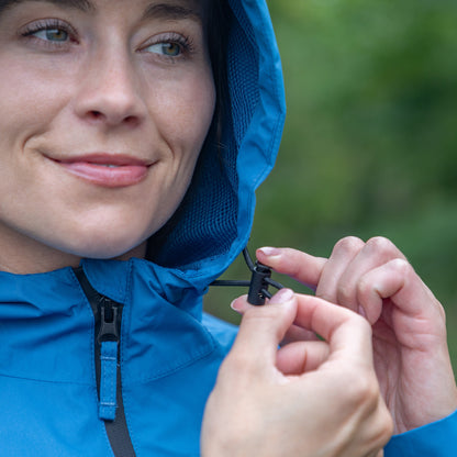 Close-up of a woman wearing a bright blue hooded jacket and adjusting the hood closure with her hands, set against a blurred green outdoor background.