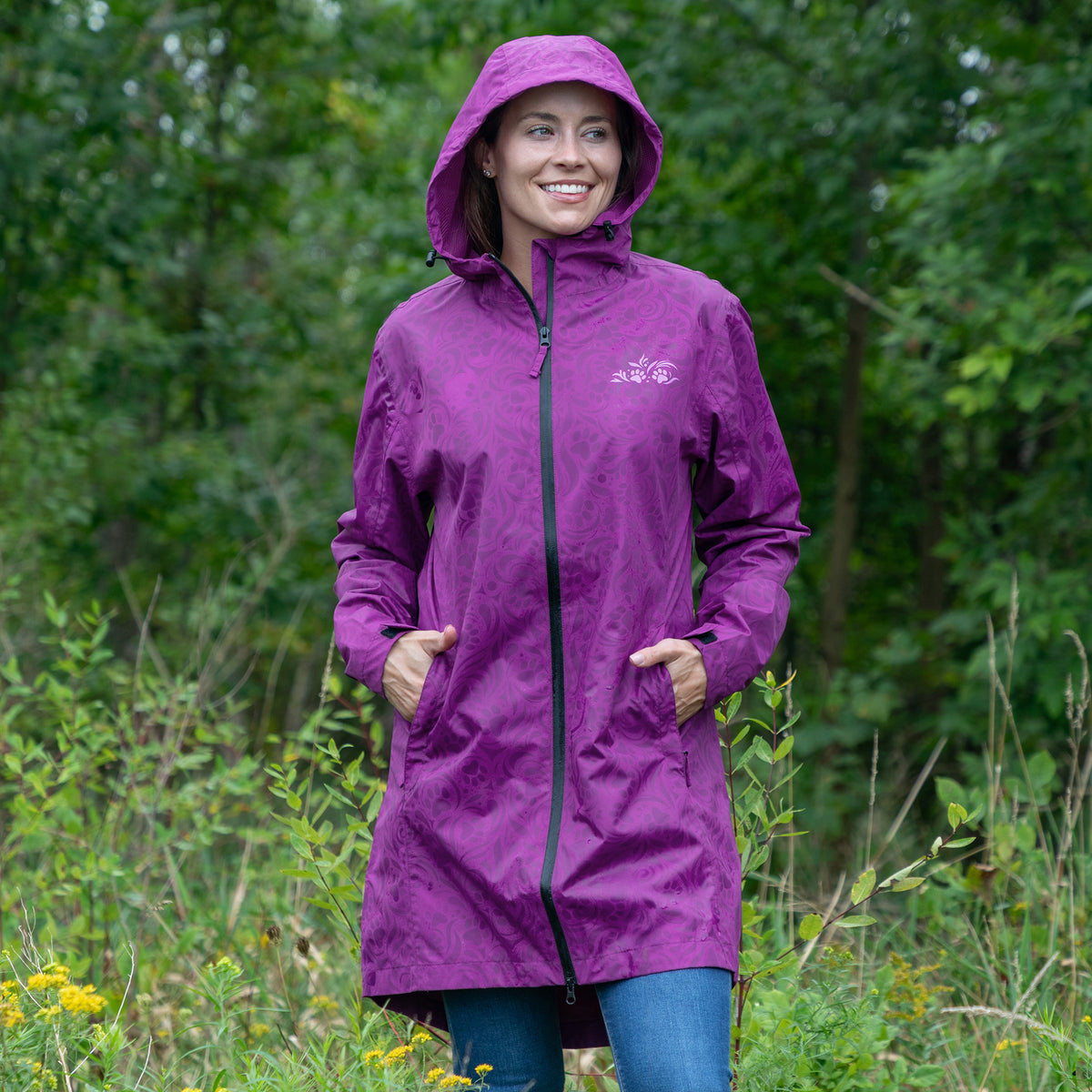 A woman wearing a purple hooded rain jacket with a black front zipper and side pockets, standing outdoors in a grassy area with green foliage behind her. The brand name 'Mossy Oak' is visible in white script on the left chest.