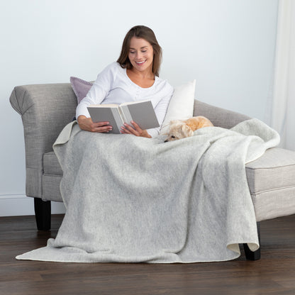 A woman sits on a light gray armchair reading a book while wrapped in a light gray blanket, with a small light-colored dog resting on the blanket. The scene is set in a bright, modern room with dark wood flooring.