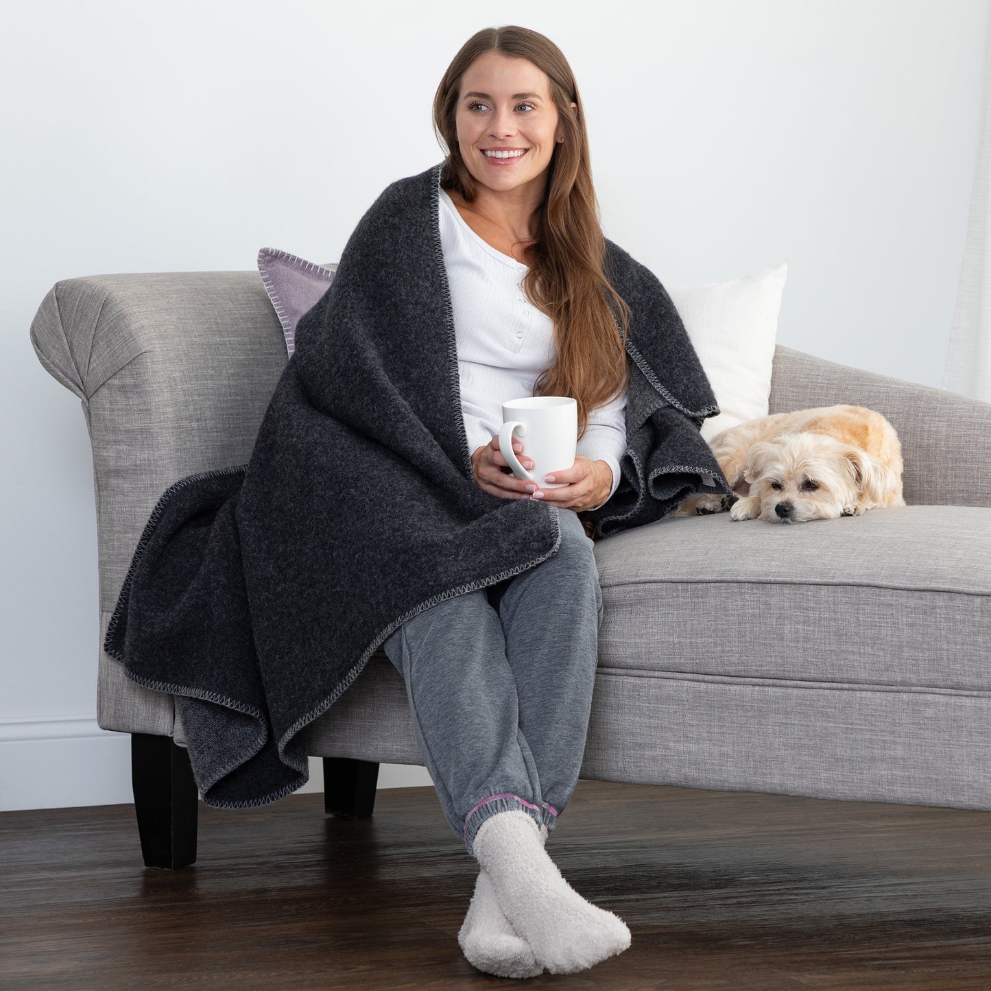 A woman sits on a light gray sofa wrapped in a dark charcoal heather throw blanket, holding a white mug, with a small cream-colored dog resting on the sofa beside her.