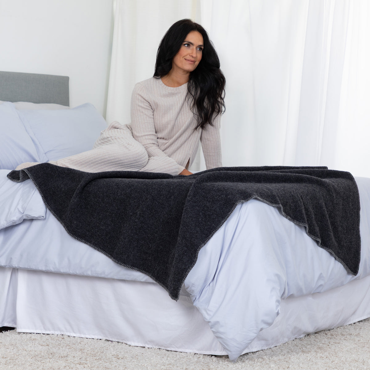 A woman sits on a bed with white bedding, draped over her lap a dark charcoal gray knit blanket or throw. She wears a light beige ribbed long-sleeve top in a bright bedroom with white curtains and a gray headboard.