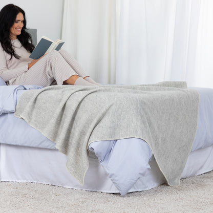 A woman in light pink pajamas sits on a bed reading a book, with a light gray heathered knit blanket draped over the bed in a bright, neutral bedroom setting.