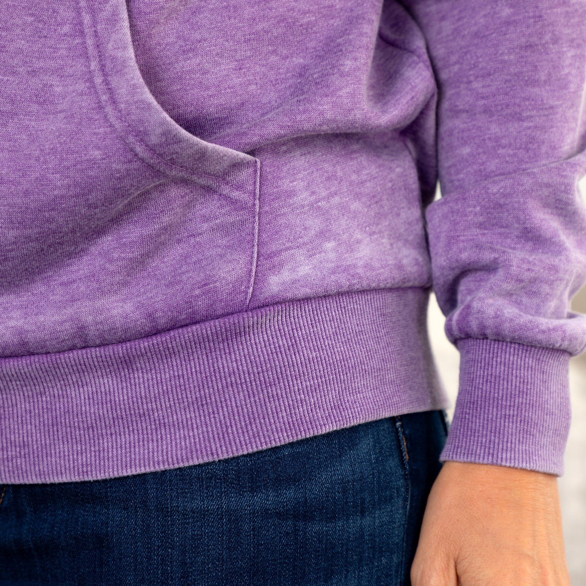 Close-up of a person wearing a heathered purple ribbed hoodie with a front kangaroo pocket and dark blue jeans.