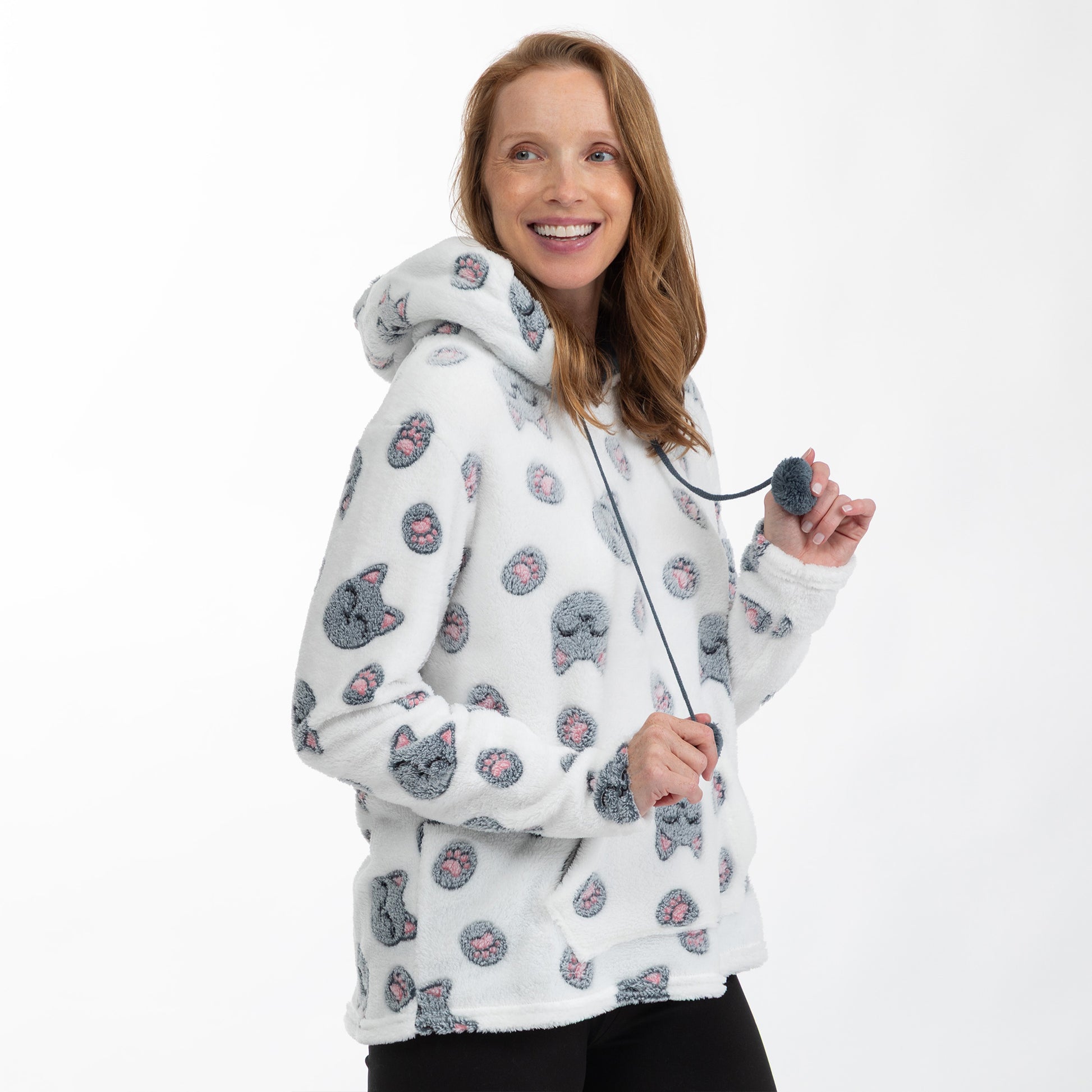 A woman wearing a white sherpa hoodie with a gray and pink cat print pattern, holding a gray pom-pom on a black cord against a plain white background.