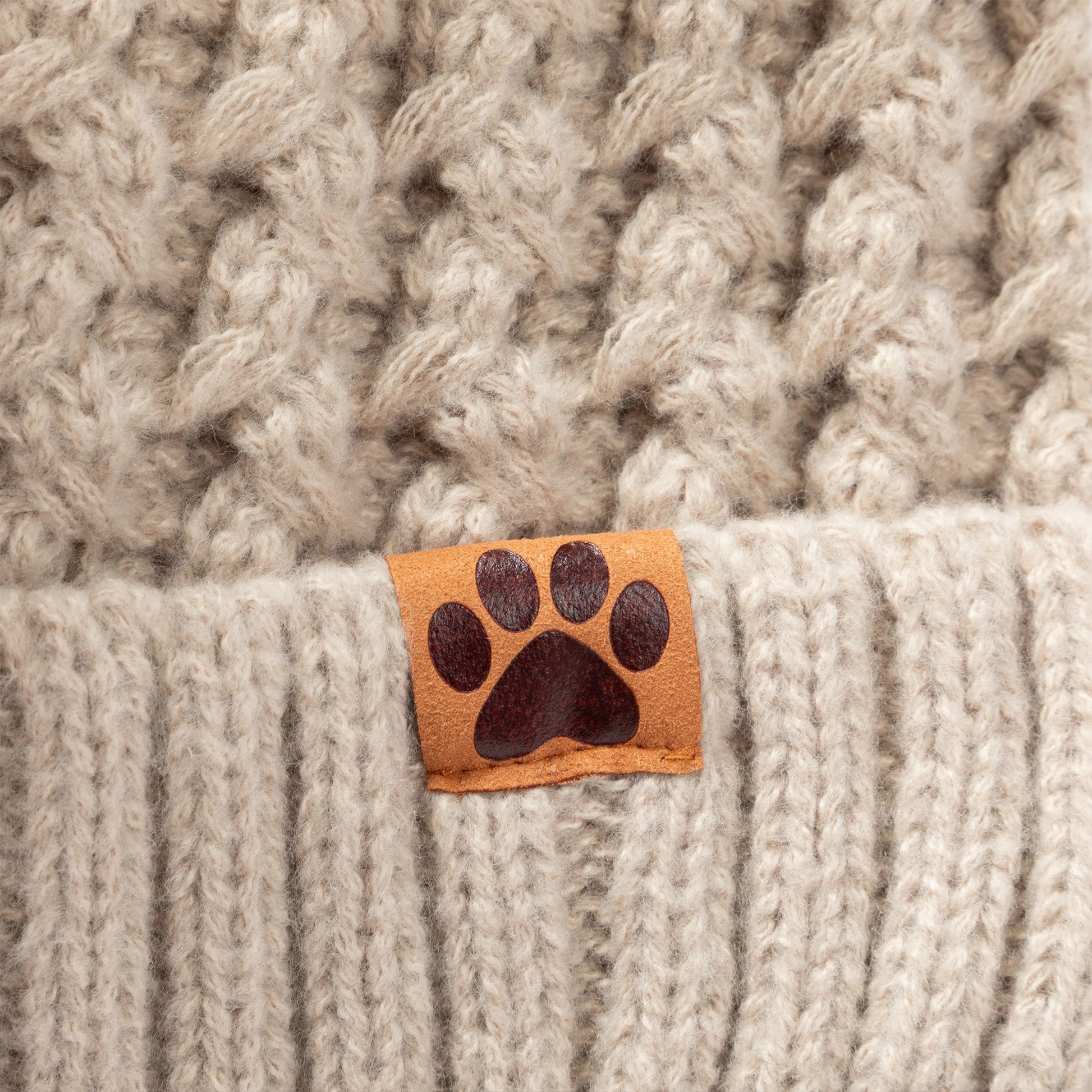 Close-up of a cream-colored chunky knit beanie with a ribbed brim and a small tan leather patch featuring a dark brown paw print.