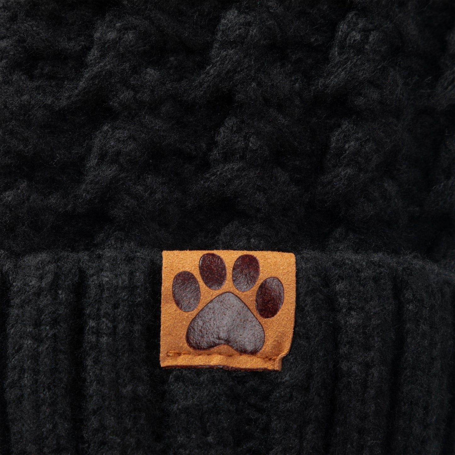 Close-up of a black textured knit beanie with a small tan square patch featuring a dark gray paw print on the front.