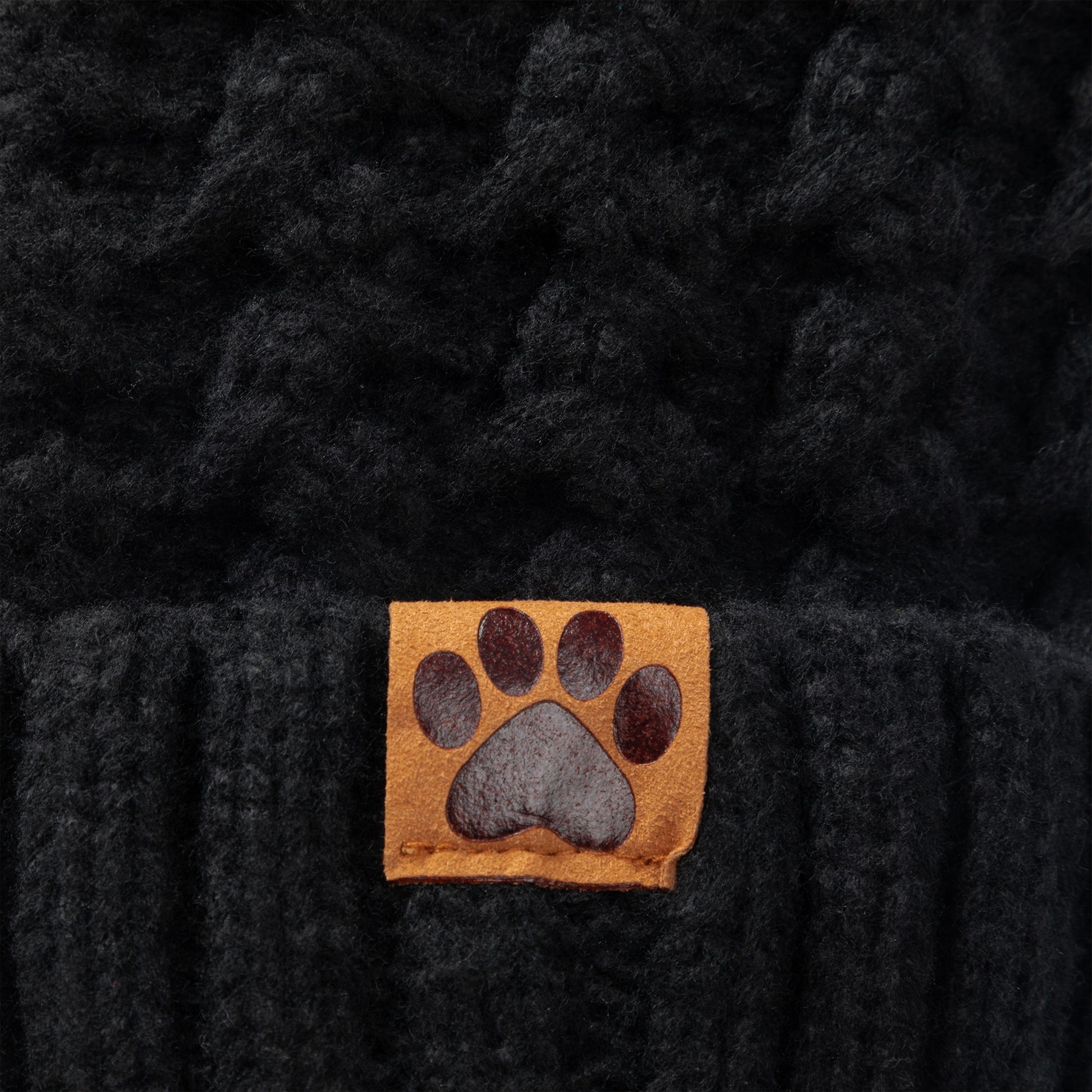 Close-up of a black textured knit beanie with a small tan square patch featuring a dark gray paw print on the front.