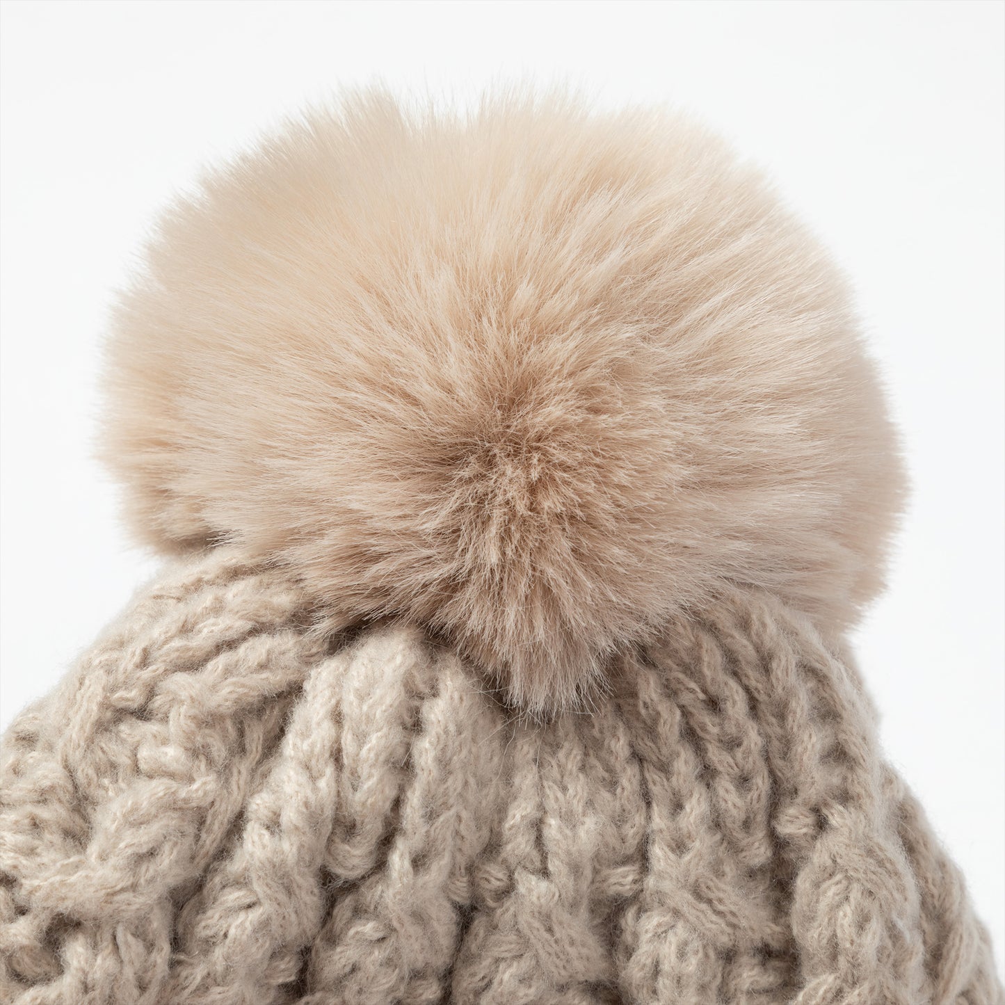 Close-up of a chunky cable-knit beanie in a warm taupe-beige color, topped with a large fluffy faux-fur pom-pom in a matching light beige tone, set against a plain white background.