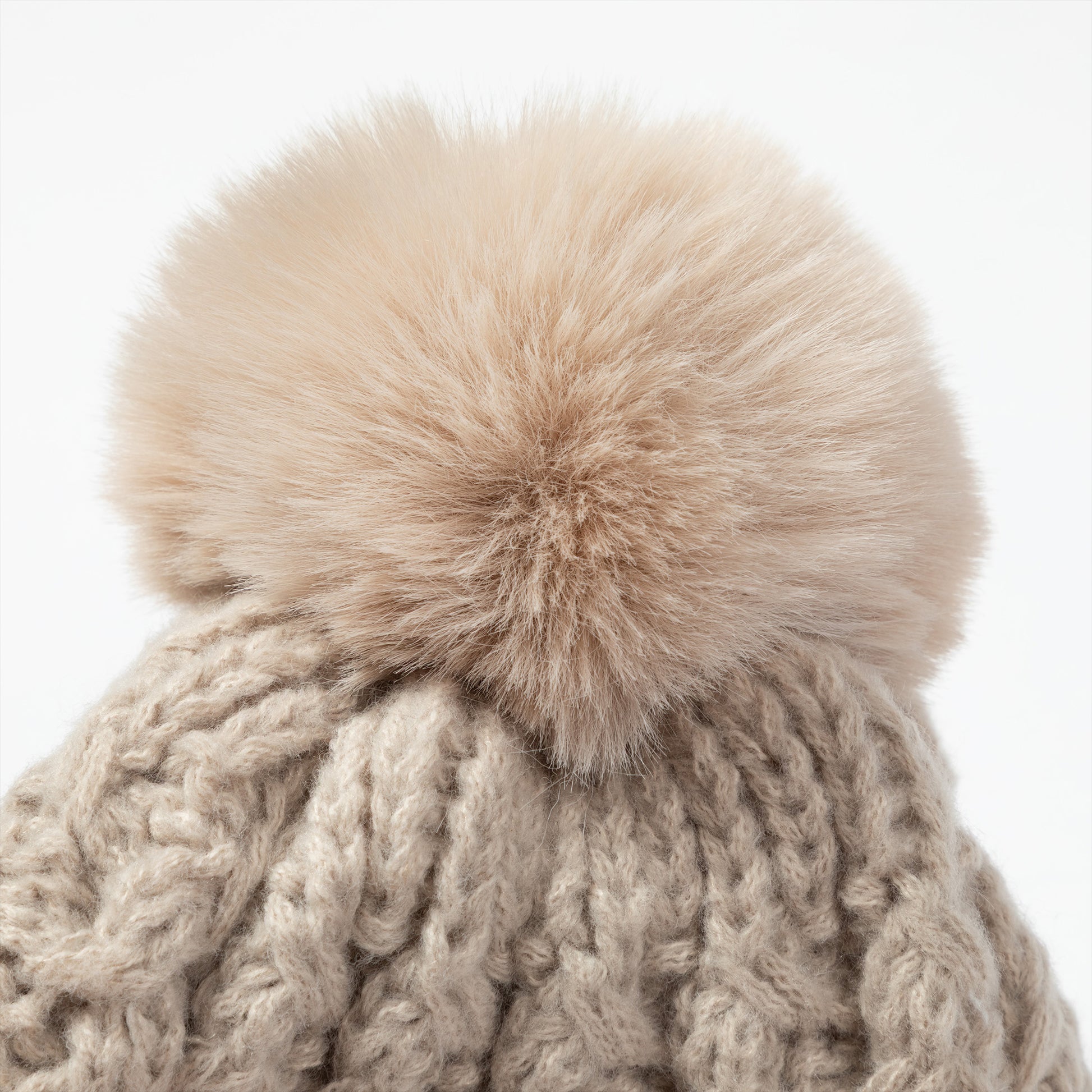 Close-up of a chunky cable-knit beanie in a warm taupe-beige color, topped with a large fluffy faux-fur pom-pom in a matching light beige tone, set against a plain white background.