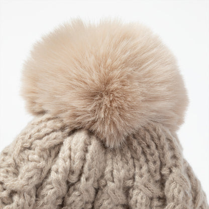 Close-up of a chunky cable-knit beanie in a warm taupe-beige color, topped with a large fluffy faux-fur pom-pom in a matching light beige tone, set against a plain white background.