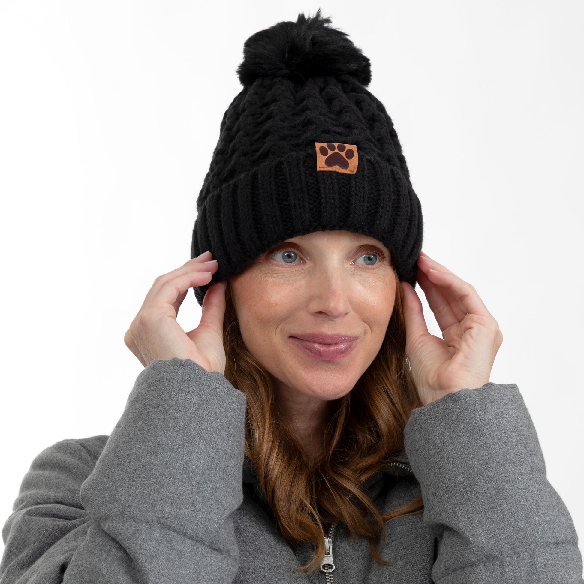 A woman wearing a black cable-knit beanie with a brown paw-print patch on the front, posing against a solid white background.