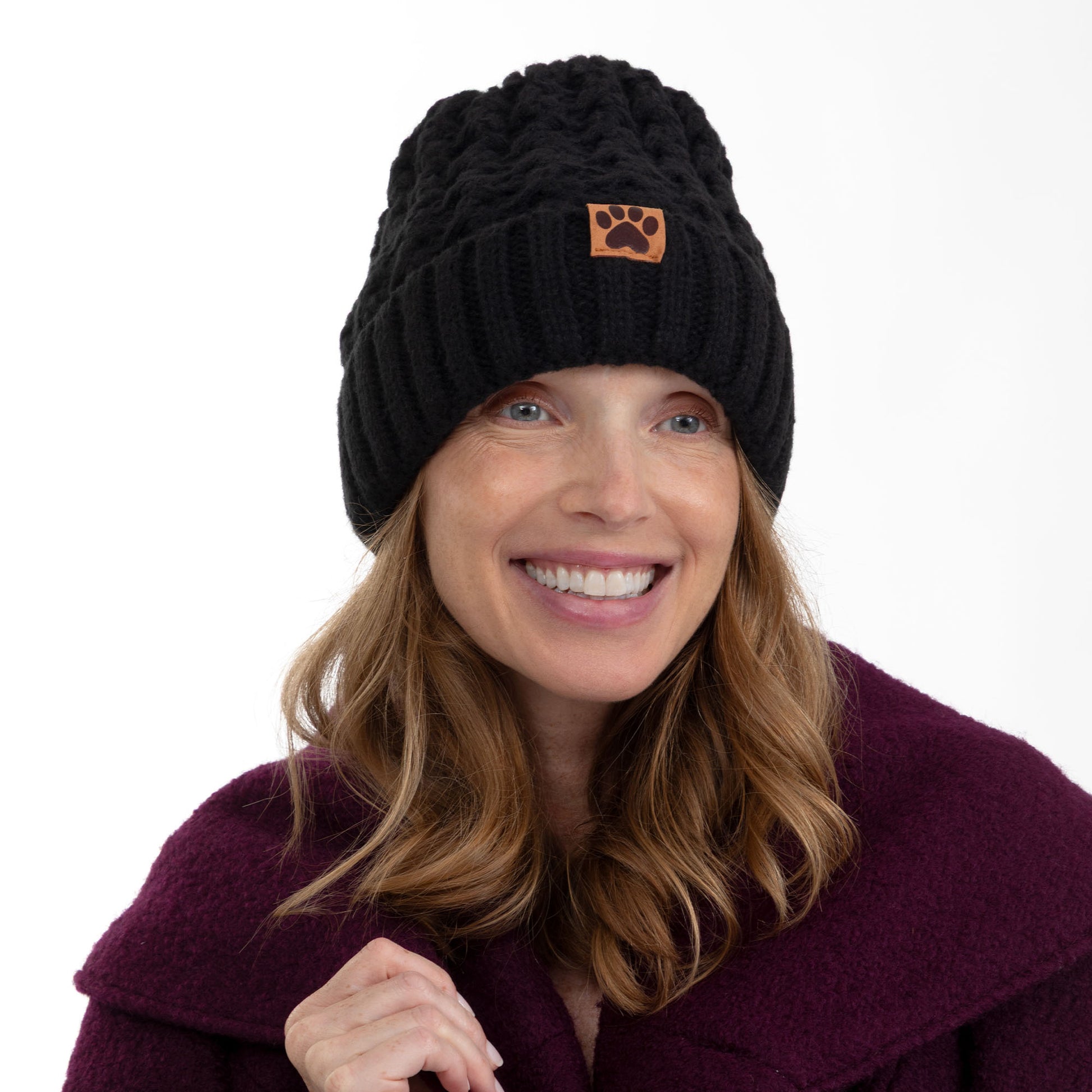 A smiling woman wearing a black cable-knit beanie with a small brown paw-print patch on the front, paired with a dark purple coat, against a plain white background.