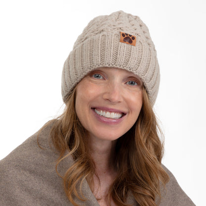 A smiling woman wearing a beige cable-knit beanie with a small brown paw-print patch on the front, against a plain white background.