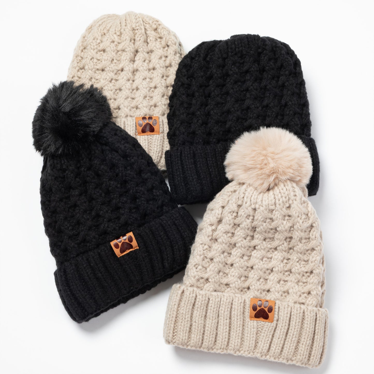 Four knit pom-pom beanies arranged on a white background, including two black and two cream/beige versions, each with a small brown paw-print patch on the cuff.