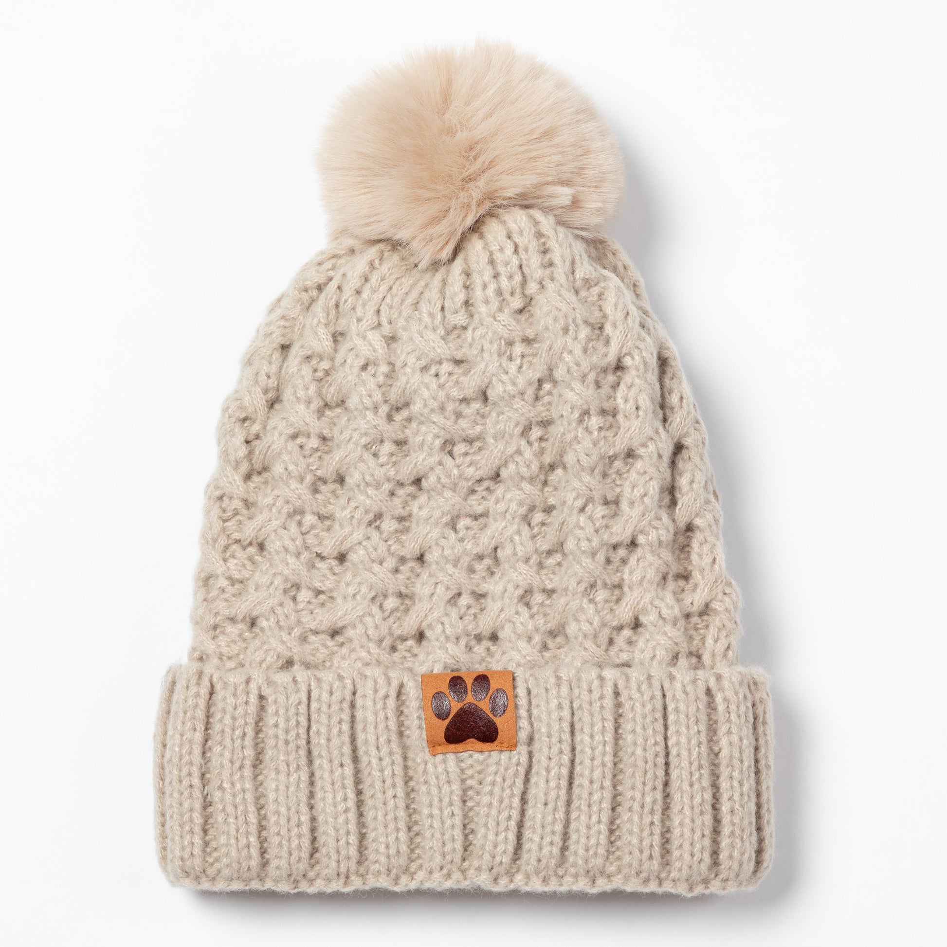 A beige cable-knit beanie hat with a matching faux-fur pom-pom on top and a folded ribbed cuff featuring a small brown paw-print patch, shown against a plain white background.