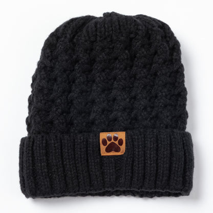 A black cable-knit beanie hat featuring a folded cuff with a small brown leather patch displaying a dark brown paw print logo, set against a plain white background.