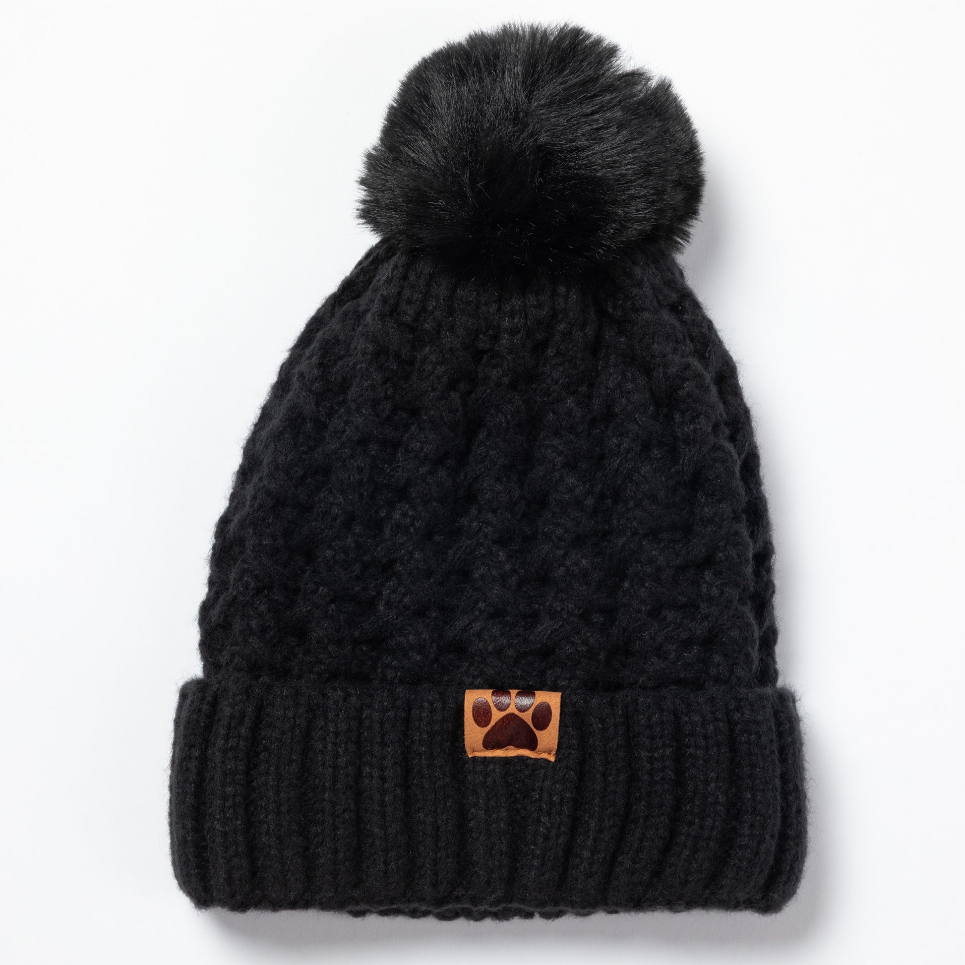 A black cable-knit beanie with a fluffy pom-pom on top and a folded ribbed cuff. A small brown leather patch on the cuff features a paw print logo, set against a clean white background.