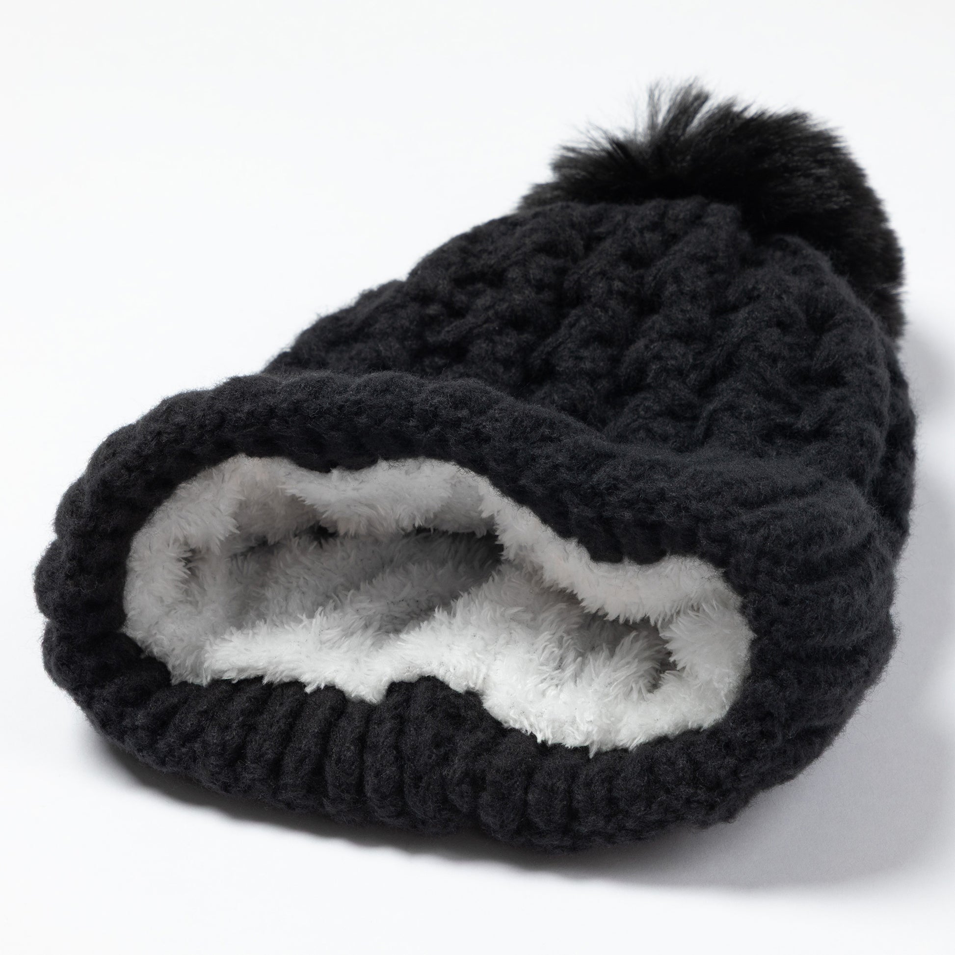 A black chunky knit beanie hat with a fluffy black pom-pom on top and a soft white faux-fur lining visible at the opening, photographed on a clean white background.