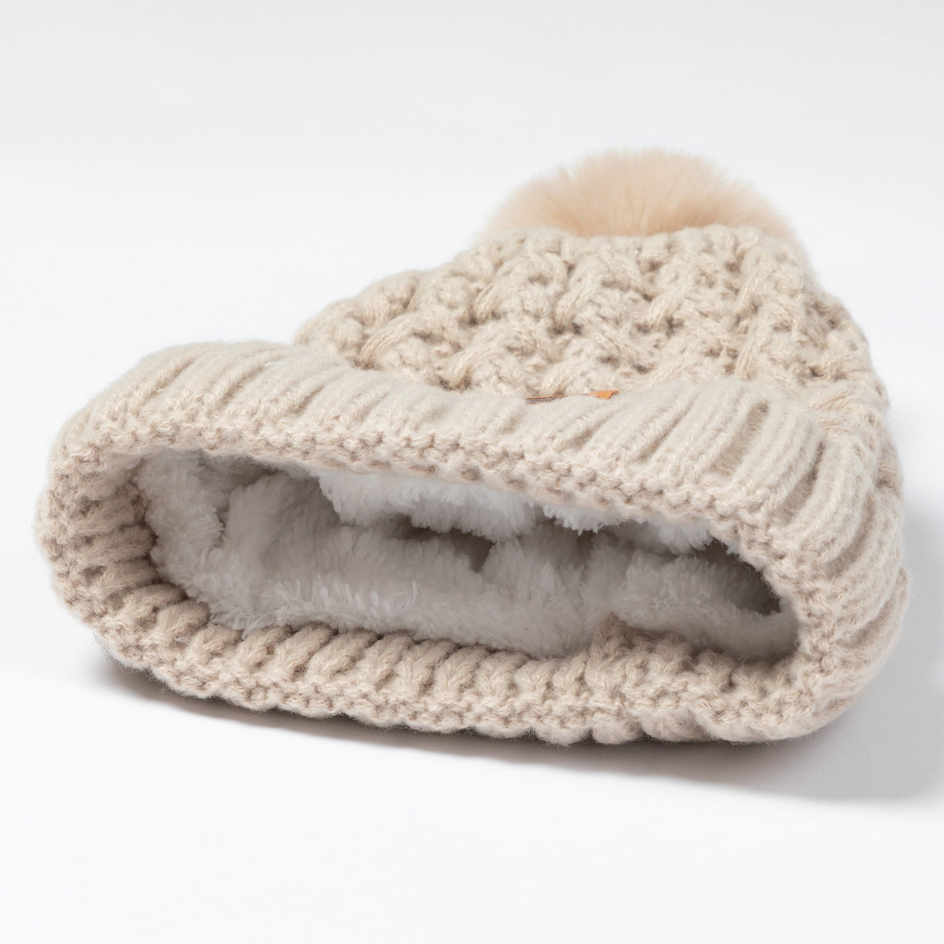 A beige chunky knit beanie hat with a fluffy pom-pom on top and a soft light gray faux-fur lining visible inside, photographed on a clean white background.