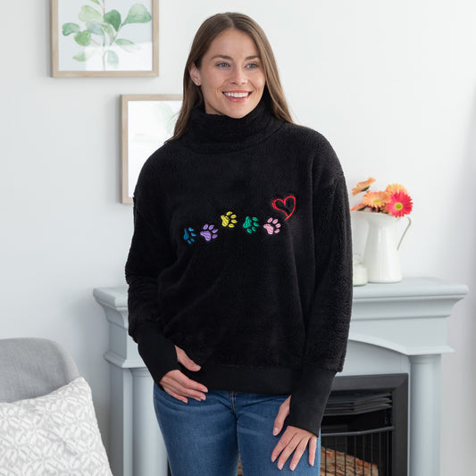 A woman wearing a black fuzzy mock neck fleece with colorful embroidered paw prints and a heart on the chest, standing in a living room in front of a fireplace.