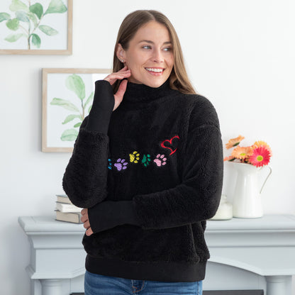 A woman wearing a black fuzzy mock neck pullover with colorful embroidered paw prints and a red heart on the front, standing in a bright room with framed botanical prints and a white mantel in the background.