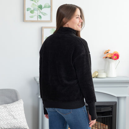 A woman wearing a black sherpa fleece pullover, shown from the back in a cozy living room setting.