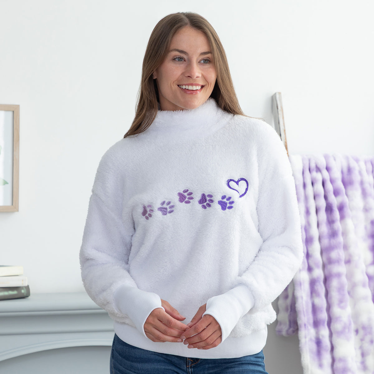 A woman wearing a white fuzzy mock-neck sweater with purple paw prints and a heart on the chest, standing in a brightly lit room.