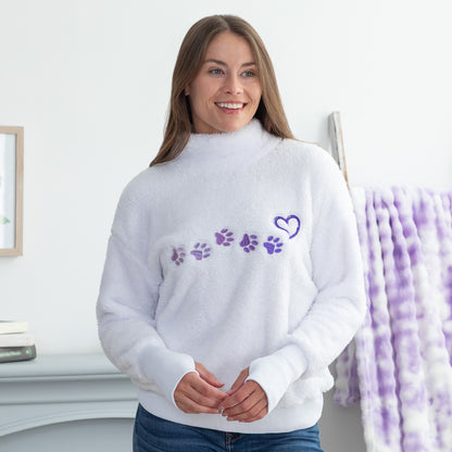 A woman wearing a white fuzzy mock-neck sweater with purple paw prints and a heart on the chest, standing in a brightly lit room.