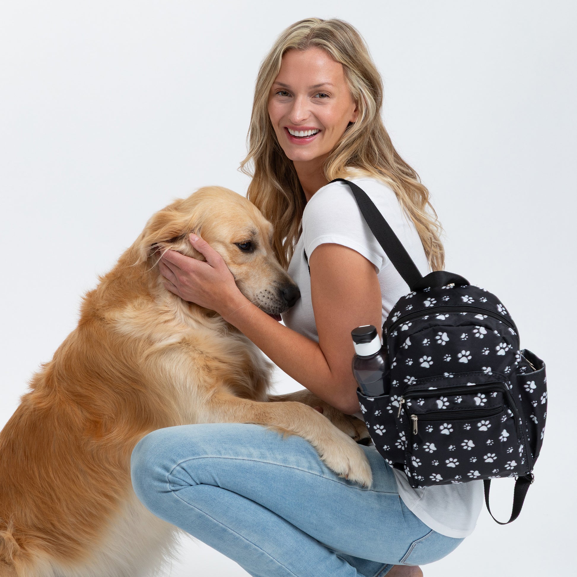 Woman petting a golden retriever while wearing a black mini backpack with white paw prints all over, against a white background.