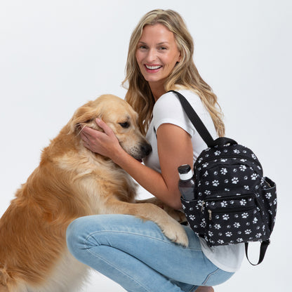 Woman petting a golden retriever while wearing a black mini backpack with white paw prints all over, against a white background.