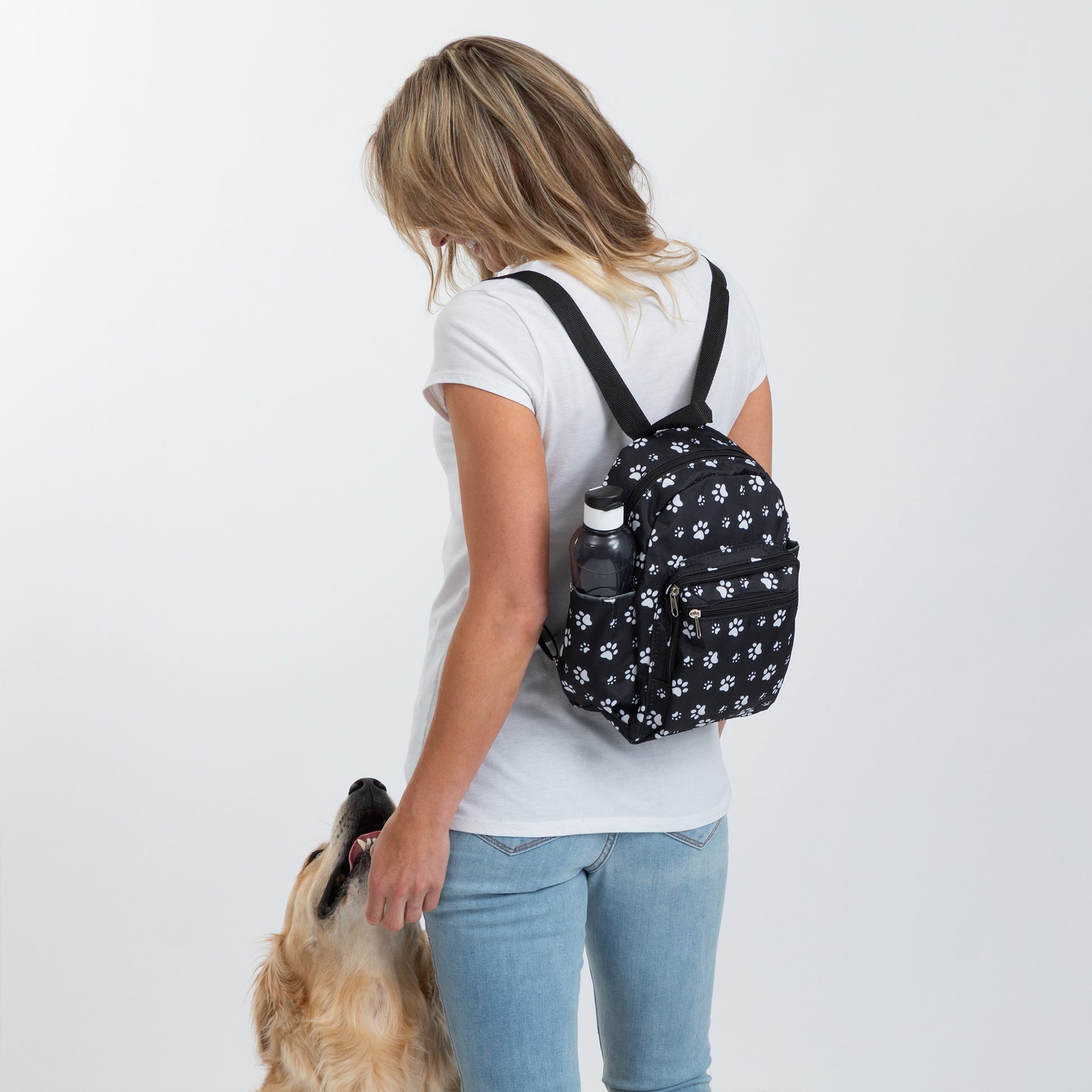 A woman wearing a white t-shirt and blue jeans is shown from behind, carrying a small black backpack with a white paw print pattern. A golden retriever dog is looking up at her.