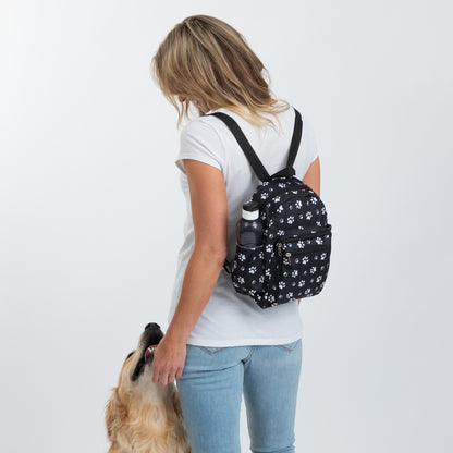 A woman wearing a white t-shirt and blue jeans is shown from behind, carrying a small black backpack with a white paw print pattern. A golden retriever dog is looking up at her.