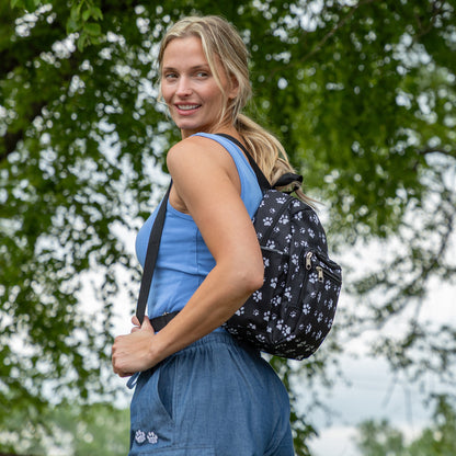 A woman outdoors wearing a black backpack with a white paw print pattern, with green trees in the background.