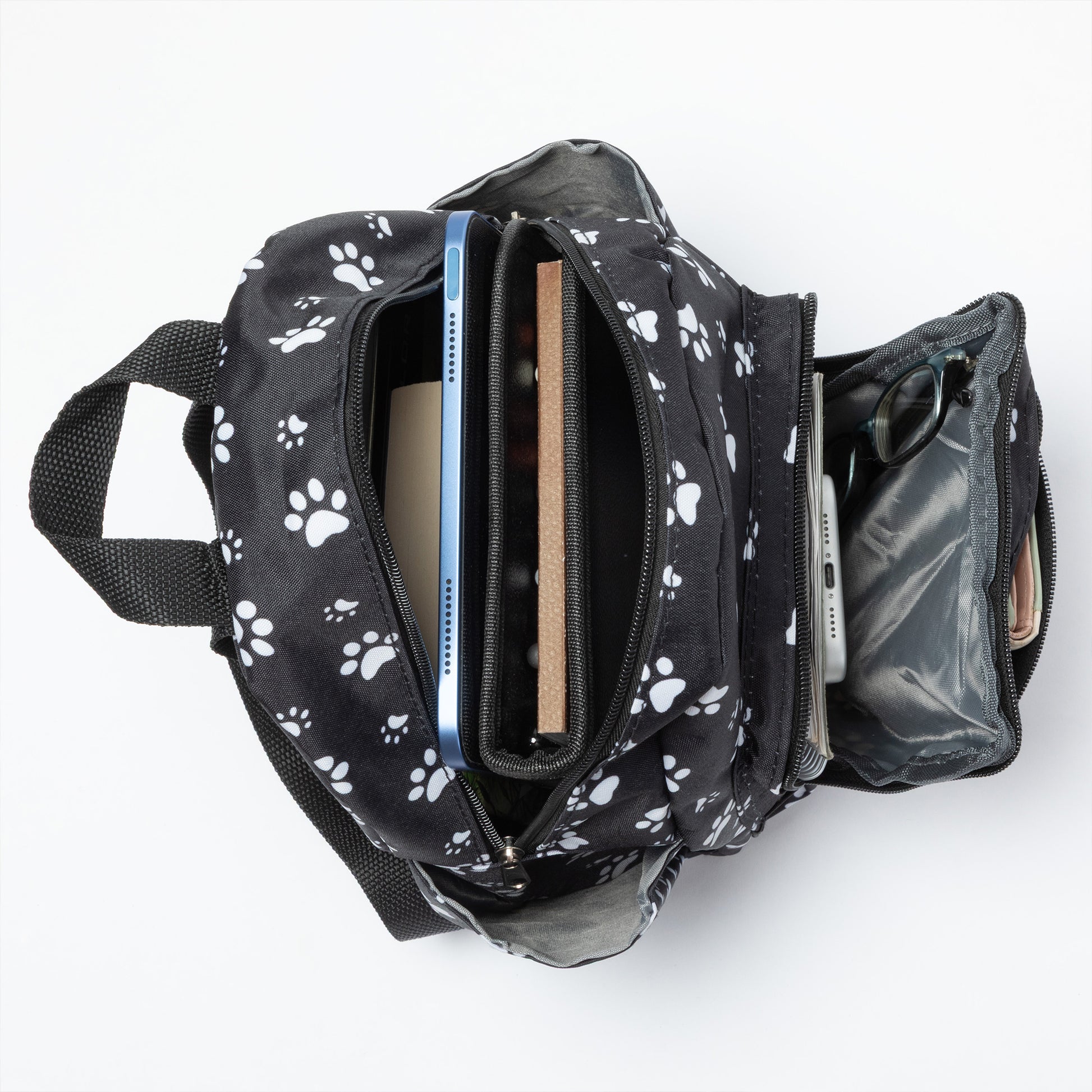 A black backpack with a white paw print pattern, shown open from a top-down view on a white background, containing a tablet, notebook, and other accessories.