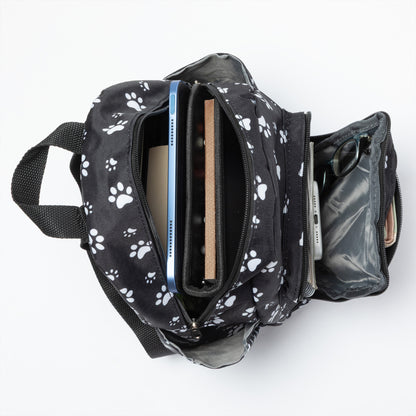 A black backpack with a white paw print pattern, shown open from a top-down view on a white background, containing a tablet, notebook, and other accessories.
