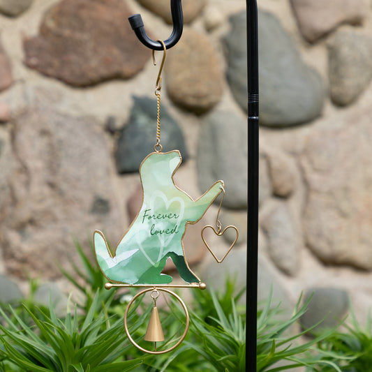 Decorative hanging wind chime with a green silhouette of a dog and text, attached to a metal hook against a stone wall background.