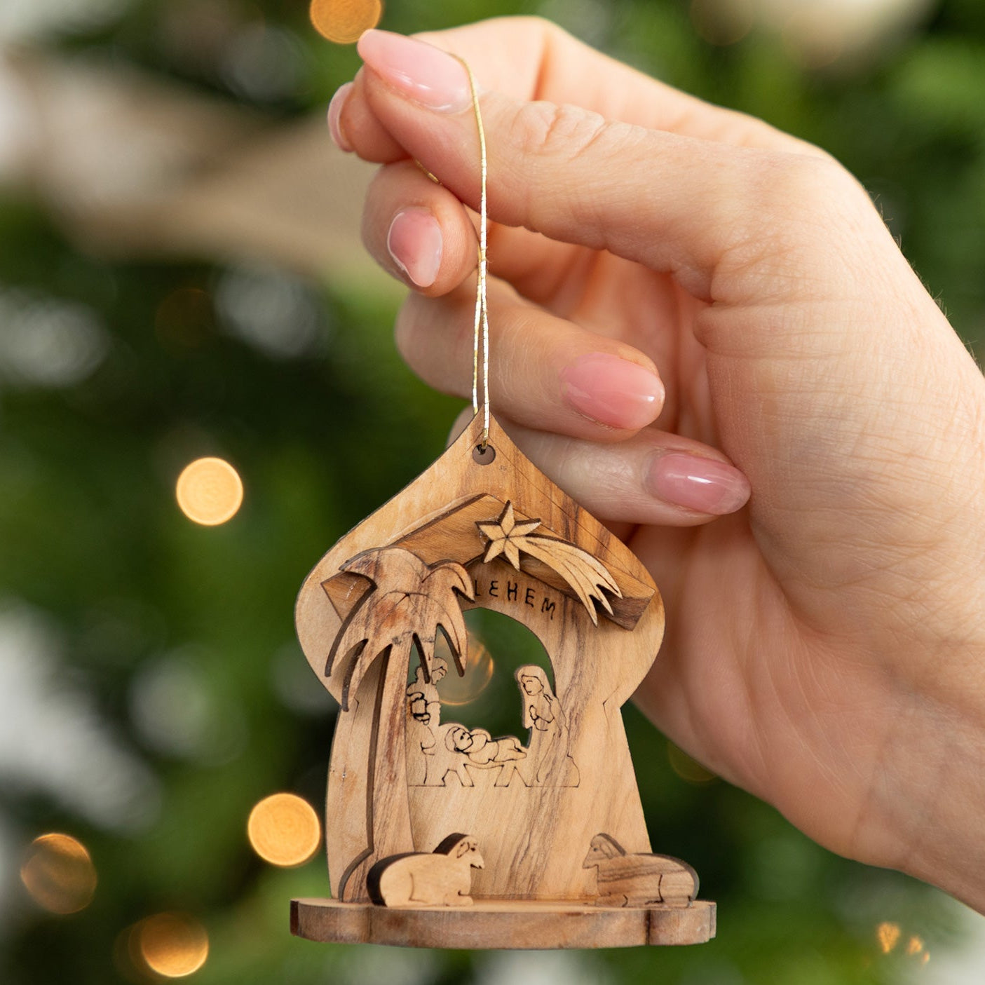 A hand holds a small carved wooden nativity ornament with a hanging string, set against a blurred Christmas tree with warm lights.