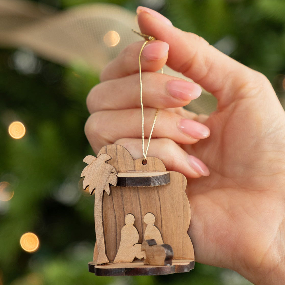 A hand holds a small wooden nativity ornament in front of a blurred Christmas tree with warm lights and silver ornaments.
