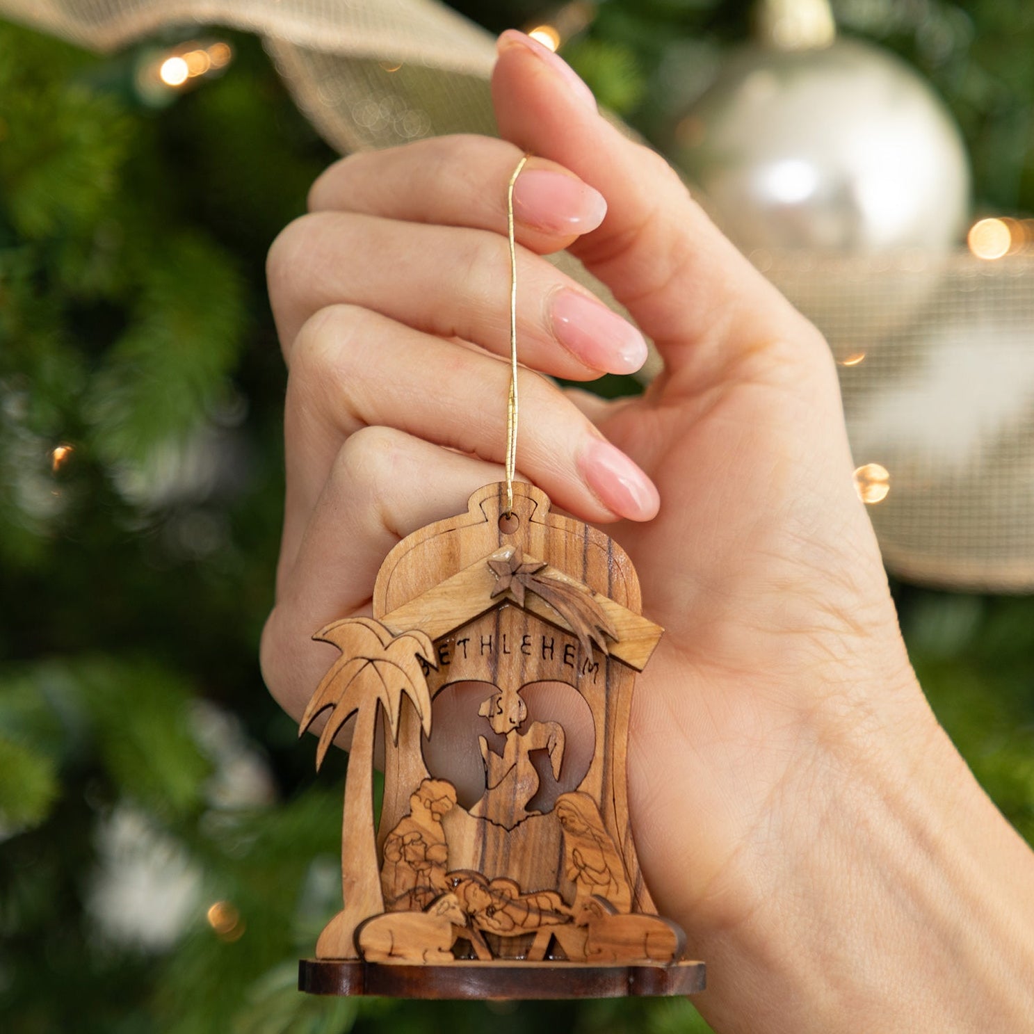 A hand holds a small wooden Christmas nativity ornament in front of a blurred Christmas tree with warm lights and ribbon.