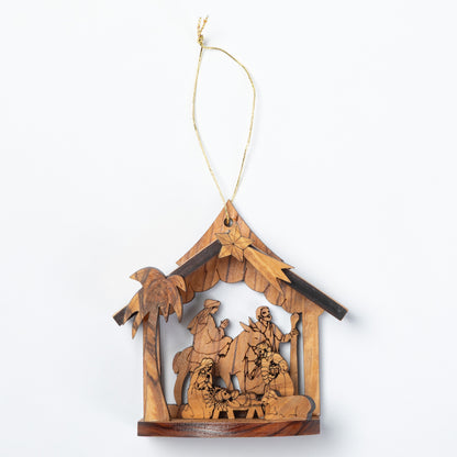A handcrafted wooden nativity scene ornament hanging from a thin string. The ornament features a house-shaped frame with a star at the peak, carved figures of the Holy Family, animals, and a palm tree, all made from light and dark natural wood tones.