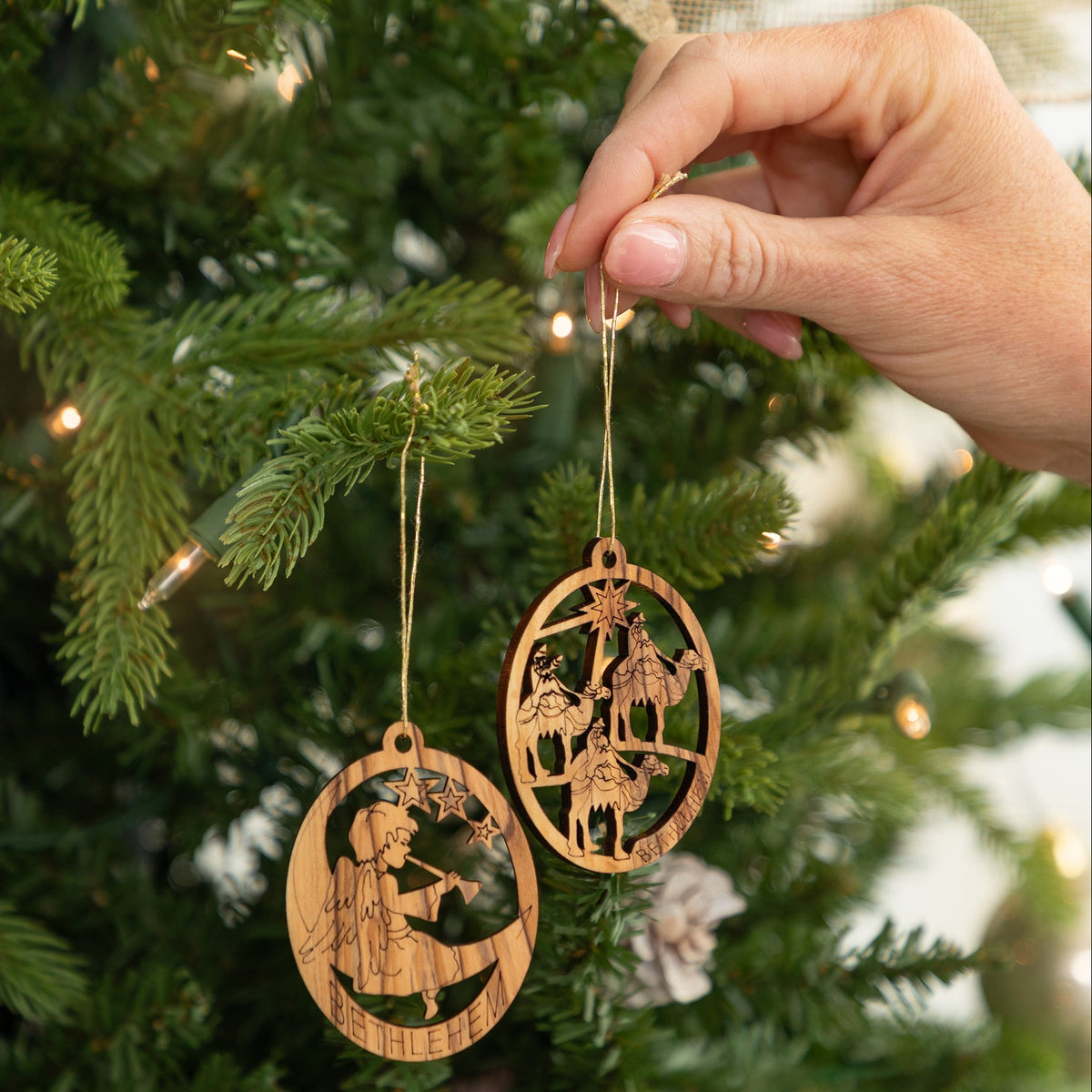 Two laser-cut wooden Christmas ornaments with detailed nativity scenes are hanging from a green Christmas tree, with a hand holding one of the ornaments.