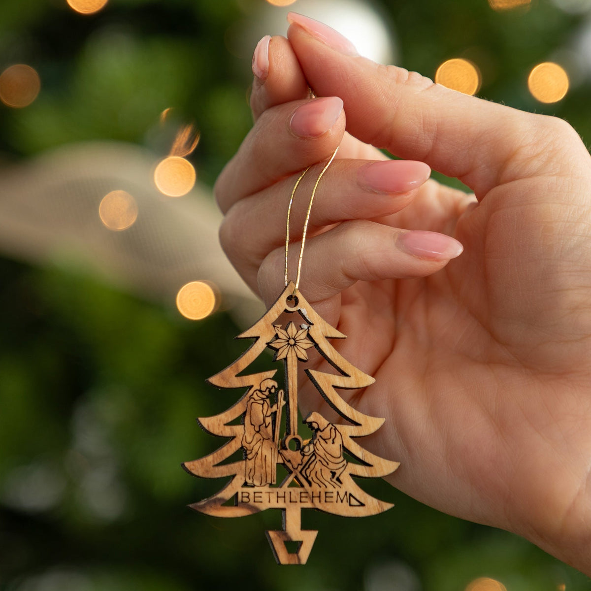 A hand holds a laser-cut wooden Christmas ornament shaped like a Christmas tree with the word 'BETHLEHEM' engraved at the bottom, set against a blurred green Christmas tree with warm bokeh lights.