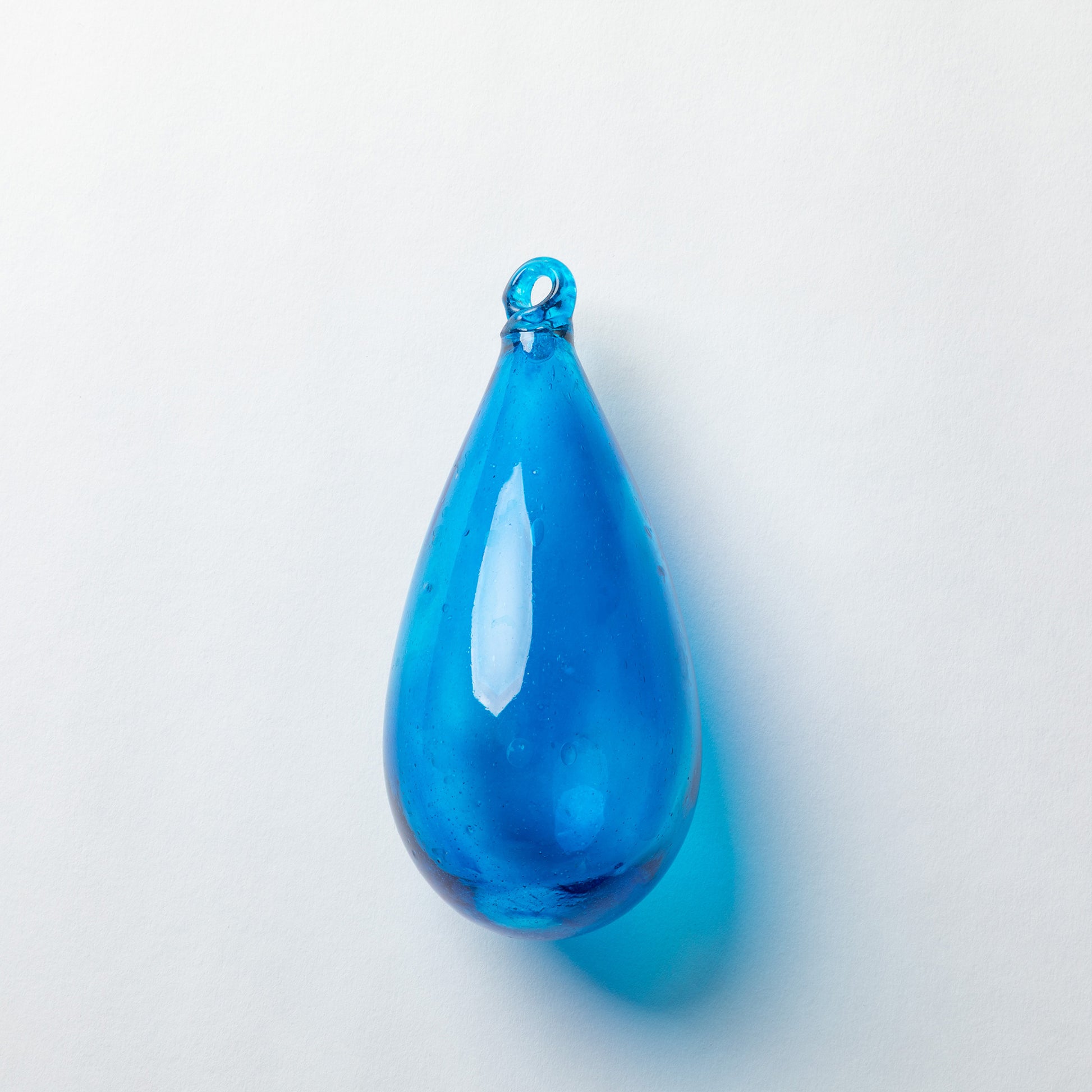 A translucent blue teardrop-shaped glass pendant with a small loop at the top, shown on a plain light gray background with a soft shadow.